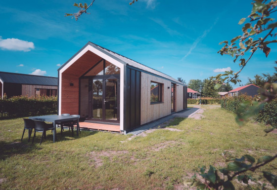 Cabaña glamping moderna con grandes ventanas y mesa exterior, rodeada de césped verde y cielo azul claro.