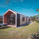 Moderne glamping hytte med store vinduer og udendørs spisebord, omgivet af grønt på solskinsdag.