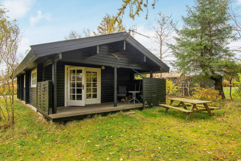 Zwarte houten cottage 'Beautiful cottage near the river' in Denemarken met overdekte veranda en tuinbank.