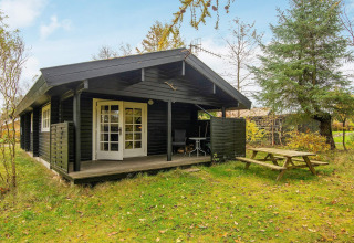Zwarte houten cottage 'Beautiful cottage near the river' in Denemarken met overdekte veranda en tuinbank.