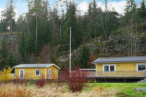 Gul feriebolig og anneks i Hällevadsholm, Sverige, omgivet af grønt skovområde og klippevæg.