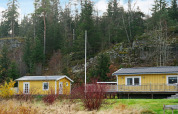 Gul feriebolig og anneks i Hällevadsholm, Sverige, omgivet af grønt skovområde og klippevæg.