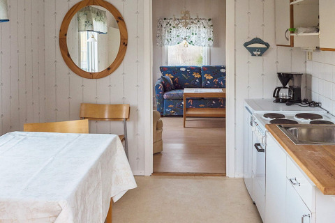 Køkken og spiseområde med bord, stole, spejl og udsigt til stue med blomsteret sofa i en svensk hytte.