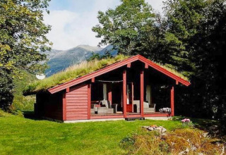 Chalet rojo de madera con techo de césped, rodeado de árboles y montañas en Olden, Dinamarca, en día soleado.