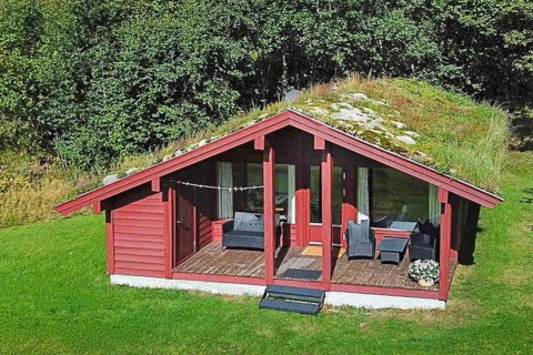 Rødt feriehus med græstag og terrasse i naturskønt område, omgivet af grønne træer og græsplæne.