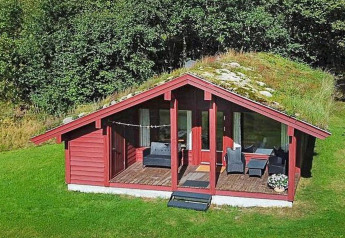 Chalet rosso con tetto erboso e veranda, immerso nel verde e circondato da alberi rigogliosi.