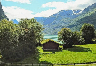 Vista desde un chalet vacacional en Olden, Dinamarca, con campos verdes, árboles, montañas y lago al fondo.