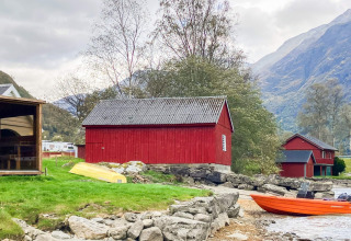 Cabaña roja de madera junto al lago con barco y montañas al fondo, casa vacacional en Olden, Dinamarca.