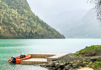 Foto de un lago con un muelle de madera, una lancha naranja y montañas al fondo en Olden.