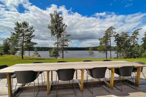 Lungo tavolo in legno con sedie nere su terrazza con vista lago e pini presso lodge in Svezia.