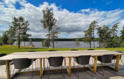 Lungo tavolo in legno con sedie nere su terrazza con vista lago e pini presso lodge in Svezia.