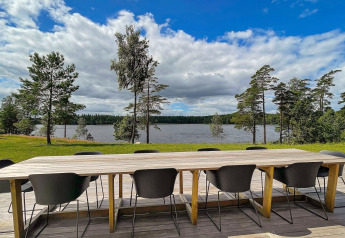 Lungo tavolo in legno con sedie nere su terrazza con vista lago e pini presso lodge in Svezia.