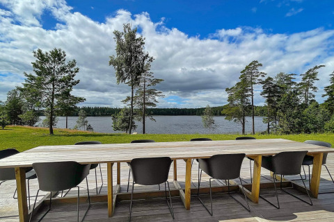 Grande table en bois avec chaises noires sur terrasse surplombant un lac et des pins en Suède.