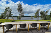 Mesa larga de madera con sillas negras en terraza, vista al lago y pinos en lodge sueco.