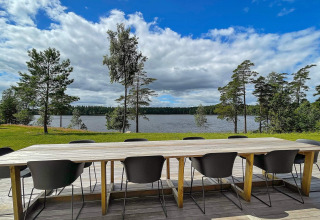 Lungo tavolo in legno con sedie nere su terrazza con vista lago e pini presso lodge in Svezia.