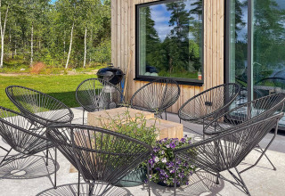Patio esterno di una villa svedese con sedie nere, tavolo in legno, fiori e sfondo verde boscoso.
