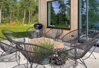 Patio esterno di una villa svedese con sedie nere, tavolo in legno, fiori e sfondo verde boscoso.