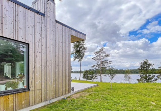 Moderne Holzvilla am Seeufer mit großen Fenstern und Blick auf Wasser und Bäume unter bewölktem Himmel.