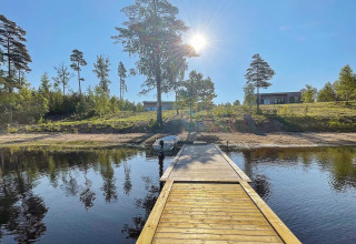 Vue ensoleillée sur un ponton en bois menant à un lac, face à deux villas modernes en Suède verdoyante.