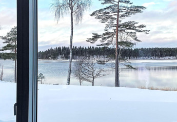Uitzicht vanuit het lodge-raam op een besneeuwd meer en dennenbomen bij Spacious Lakeside Villa with in Zweden.