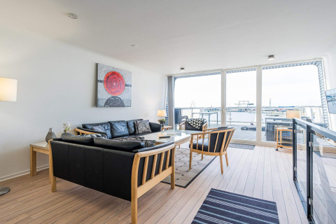 Woonkamer op een woonboot met zwart lederen bank, grote ramen en havenzicht in Hemmet, Denemarken.
