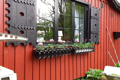 Sort vinduesramme og udskårne skodder på en rød træhytte med blomster foran, Sverige, Tjörnarp.