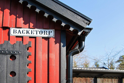 Rød træhytte med sort tag og et skilt med teksten 'BACKETORP' ved feriebolig i Tjörnarp, Sverige.