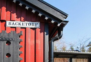 Rood houten chalet met zwart dak en bord 'BACKETORP' bij vakantiehuis in Tjörnarp, Zweden.