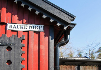 Cabane en bois rouge avec toit noir et panneau 'BACKETORP' près d'un gîte à Tjörnarp, Suède.