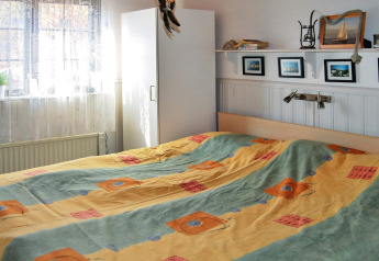 Schlafzimmer mit buntem Bettbezug, Fenster mit Gardinen, weißem Schrank und Wandregal in einer Hütte.