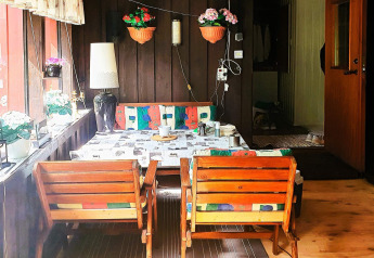 Coin repas chaleureux en cabane avec chaises en bois, coussins colorés et fleurs en pots à la fenêtre.