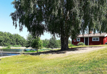 Ferienhaus in Norrtälje, Schweden, mit rotem Haus, See, Badesteg und grünem Ufer bei Sonne.