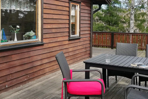 Terraza de madera con muebles negros, cojín rosa brillante y vista al bosque, en casa de vacaciones en Norrtälje.