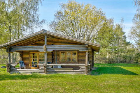 Træhytte med veranda i Eskebjerg, Danmark. Omgivet af grøn plæne og træer under en blå himmel.