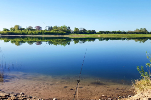 Vista tranquila de un lago en Eskebjerg, Dinamarca, con reflejo de árboles y caña de pescar en primer plano.