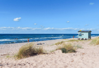 Sandstrand med marehalm, folk i vandet og et livredderhus ved havet, Melby, Danmark, på en solskinsdag.