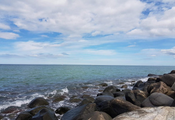 Kystudsigt fra en feriebolig i Melby, Danmark, med bølger, store sten og delvist skyet himmel.