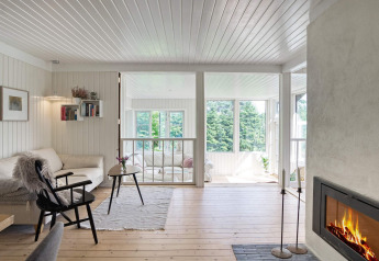 Wohnzimmer mit Kamin, hellen Holzwänden und großen Fenstern mit Blick ins Grüne im Melby, Dänemark.
