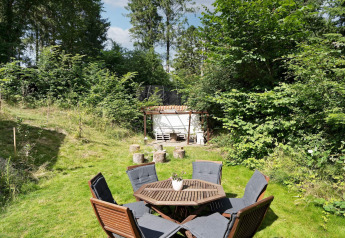 Udendørs siddeområde med bord, stole og havehytte omgivet af grøn natur ved feriehus i Melby, Danmark.