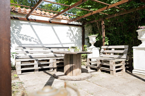 Espace extérieur avec bancs en palettes et table touret sous pergola, entouré de verdure et de soleil.