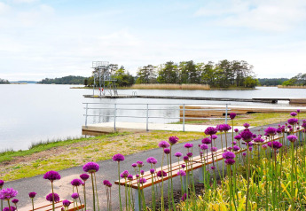 Vista al lago con plataforma de buceo, muelles de madera y flores moradas en casa de vacaciones en Mönsterås, Suecia.