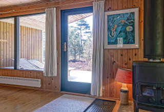 Interior de una cabaña con paredes de madera, chimenea, cuadro y vista a la naturaleza en Snogebaek, Dinamarca.