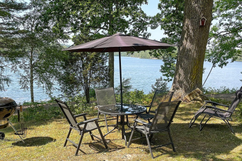 Gartenmöbel mit Sonnenschirm am Seeufer, umgeben von Bäumen, bei einer Ferienhütte in Rimforsa, Schweden.