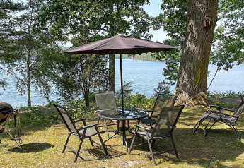Gartenmöbel mit Sonnenschirm am Seeufer, umgeben von Bäumen, bei einer Ferienhütte in Rimforsa, Schweden.