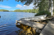 Ruderboot am Seeufer bei Ferienhaus in Rimforsa, Schweden, mit Steg, Bäumen und klarem Himmel.