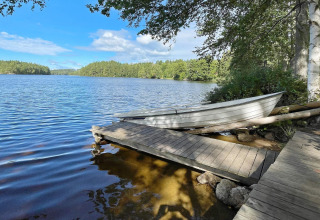 Træbåd på søbred ved feriebolig i Rimforsa, Sverige, med træbro, blå himmel og skov i baggrunden.