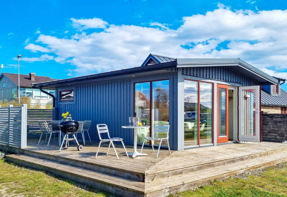 Blaues Ferienhaus mit Holzterrasse, Gartenmöbeln und Grill unter blauem Himmel in Löttorp, Schweden.