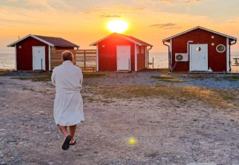 Personne en peignoir marche vers trois cabanes rouges au bord de la mer au coucher du soleil à Löttorp, Suède.