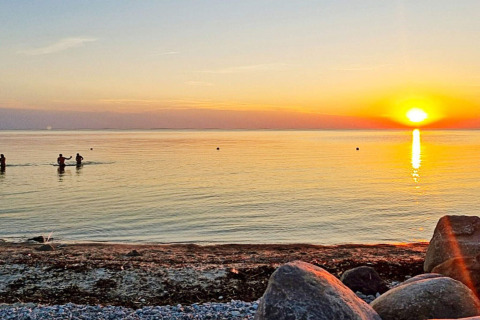 Solnedgang ved stranden i Löttorp, Sverige, med folk i vandet og sten på kysten nær en hytte.