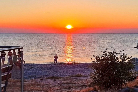 Solnedgang over havet ved stranden nær Löttorp, Sverige, med en person og busk i forgrunden.